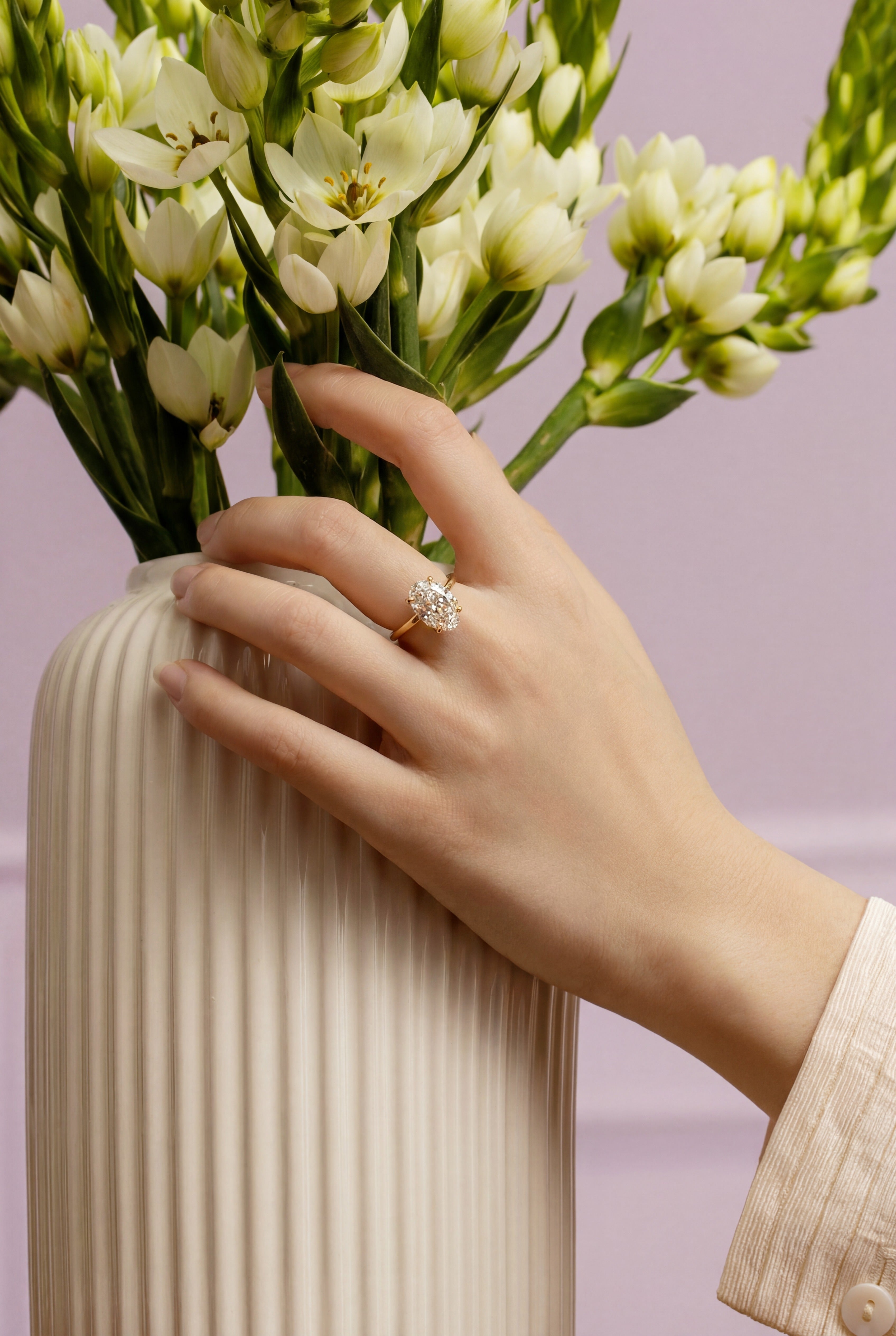 Hand holding a vase with flowers against a light purple background, main focus on the 14k solid yellow gold lab grown diamond.
